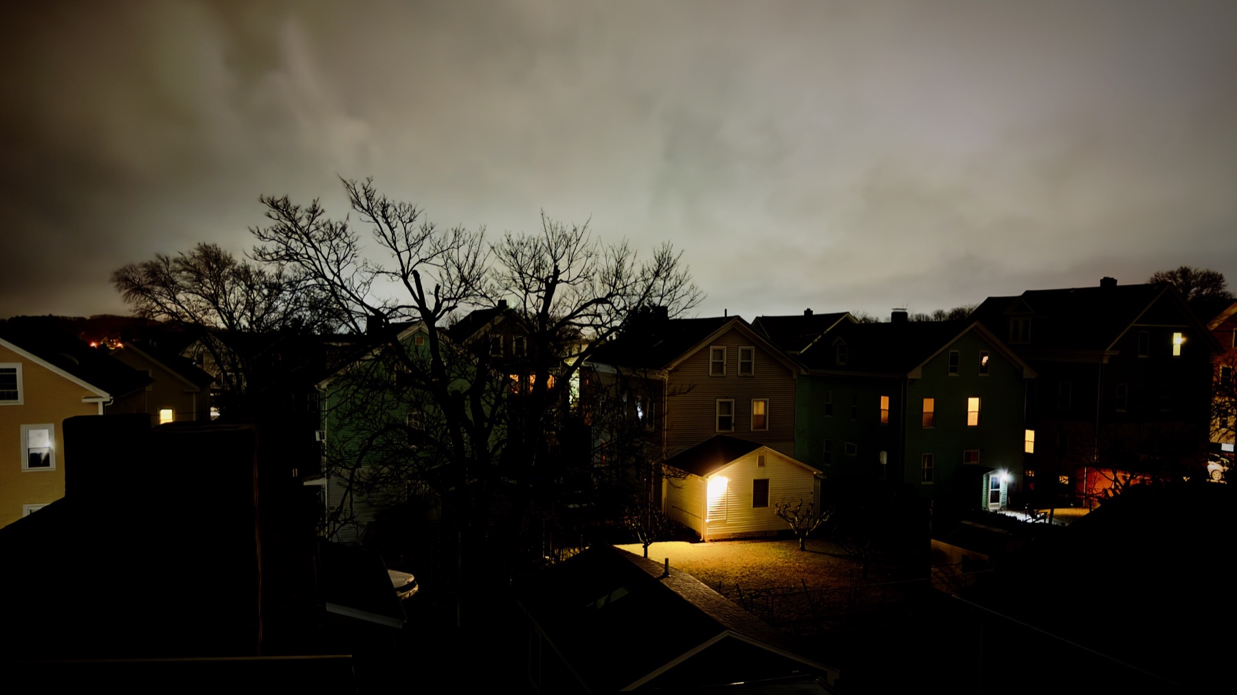 Wood-frame houses at night under an overcast sky, one backyard lit by a single porch light